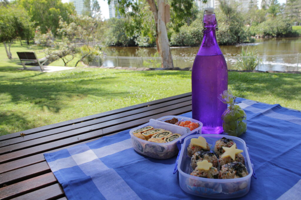 purple glass water bottle and 2 plastic lunch boxes with finger foods on a blue towel spread on a wooden picnic table at a riverfront park